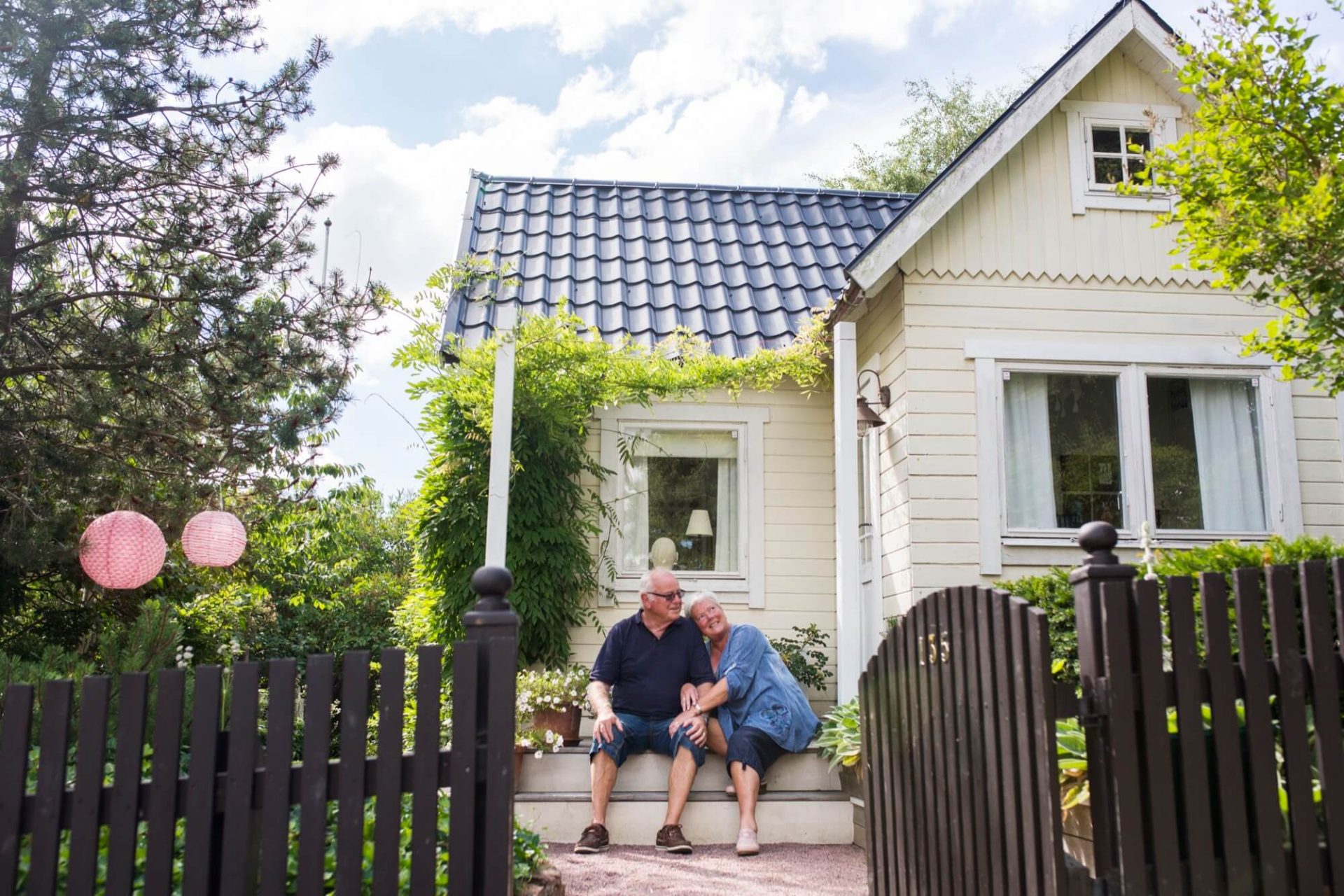 Ett solgult trähus täckt av ett mörkt tak, inbäddat i grönt, där ett äldre par avnjuter sin tid på verandan